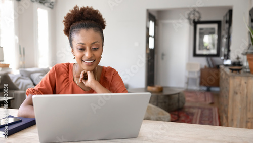 Positive focused young African American freelance professional woman working at laptop at home office workplace, touching chin, watching content, talking on video conference call. Banner shot