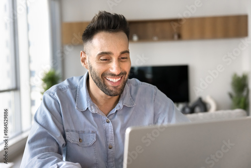 Smiling man using laptop seated at table at home office, engaged in online meeting or webinar, watching content or websurfing, learning new business or amusing platform on weekend at home. AI, tech