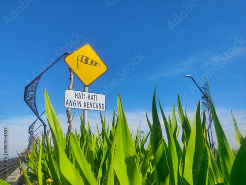 A yellow warning sign indicating strong wind conditions stands above tall green plants against a bright blue sky, featuring Indonesian text that reads “Hati-hati Angin Kencang” (Caution: Strong Winds)
