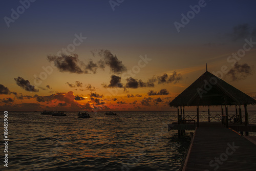 Serene sunset over ocean with wooden pier and thatched roof gazebo, silhouetted against vibrant sky. Boats are visible on water, adding to tranquil scene