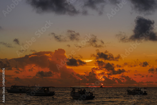  Sunset over ocean with boats silhouetted against vibrant orange and purple sky, creating tranquil and picturesque seascape