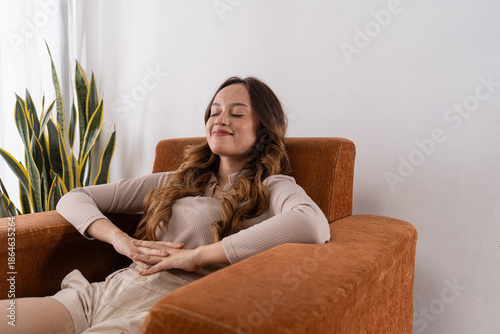 Caucasian woman reclining in armchair with hands resting on stomach, eyes closed and serene expression, warm textured upholstery