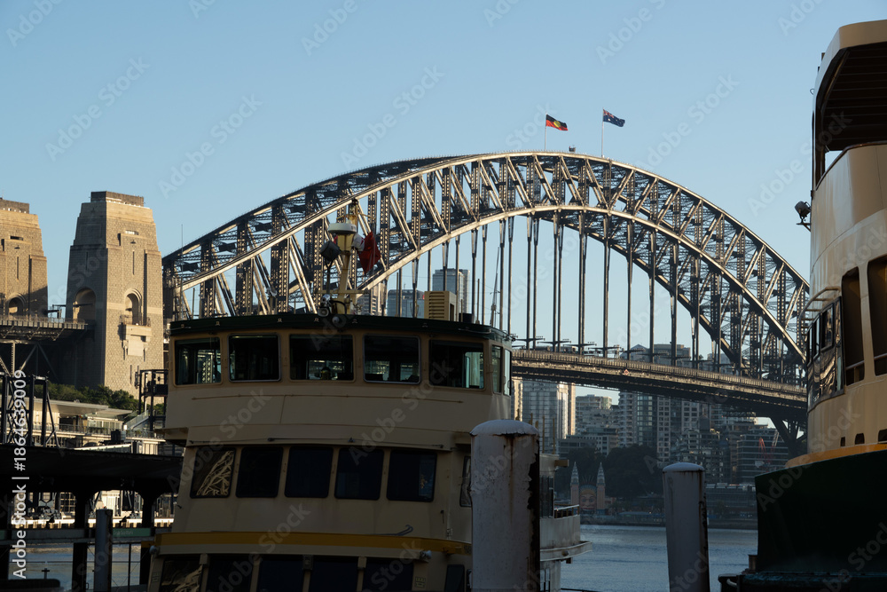 Fototapeta premium Iconic Sydney Harbour Bridge and Massive Pylon Towers Framed by a Classic Ferry Under a Clear Blue Morning Sky