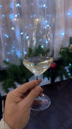 Man's point of view holding a wine glass in hand, gazing at a cozy New Year's interior with twinkling lights, decorated tree, and festive decor glowing warmly.
