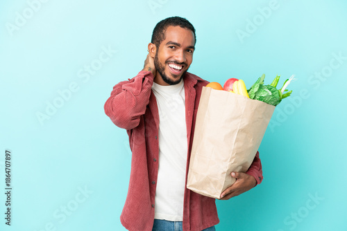 Young latin man holding a grocery shopping bag isolated on blue background laughing