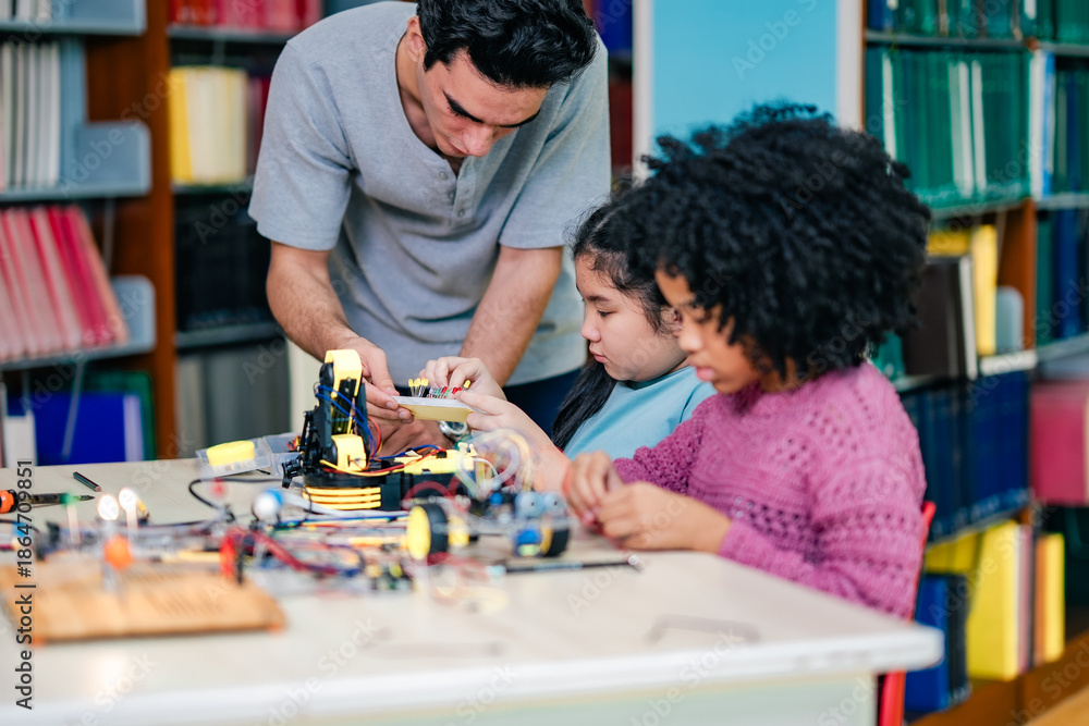 Fototapeta premium Teacher assists children during robotics build session, sharing technical knowledge, electronics practice, STEM learning experience, cooperative classroom interaction, guided education moment.