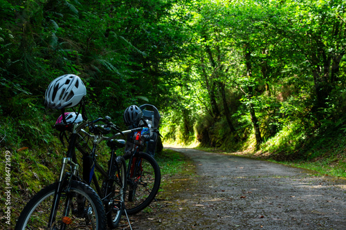 Two bikes are parked on a dirt road in a forest. The bikes are both black and have helmets on them. The helmets are placed on the handlebars of the bikes. The bikes are parked next to each other
