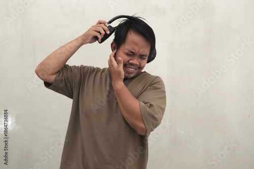 Southeast Asian man wearing casual t-shirt and headphones reacting with earache and discomfort, highlighting hearing problems, loud sound issues, and poor audio experience for health