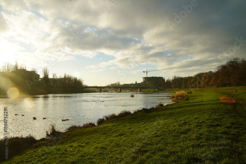 Panoramic view of a wide river in an urban park. Soft golden sunset light illuminates the grassy riverbank while dramatic clouds hang over the distant city buildings and cranes.
