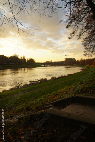 Vertical shot of a bridge crossing a river with the bright evening sun directly above the water. Strong backlight turns the bridge and riverbank into silhouettes while the surface of the water glows