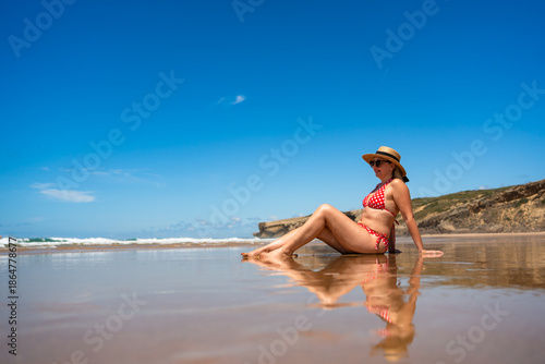 Beautiful middle-aged woman model sitting on sandy beach in summertime. Side view. Monte Clerigo beach on Algarve coast in Portugal