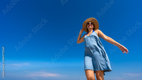 Portrait of beautiful happy middle-aged woman in summer dress and sun hat standing against background of blue sky in summertime. Front view