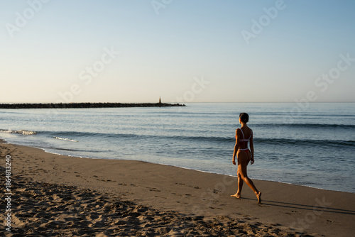 Fit young woman walking barefoot on sandy beach during sunrise, wearing red bikini, looking ahead with calm expression, gentle ocean waves in background under soft early morning light