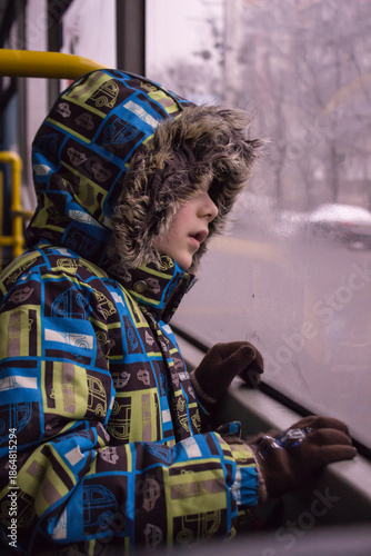 Little child looking through the window: a boy in a bright winter jacket with a faux fur hood and gloves. 
