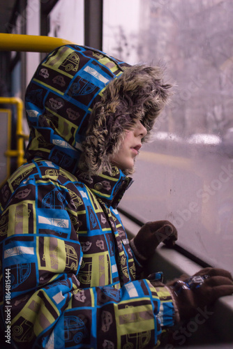 Little child looking through the window: a boy in a bright winter jacket with a faux fur hood and gloves. 