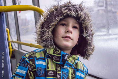 Little child looking through the window: a boy in a bright winter jacket with a faux fur hood and gloves. 