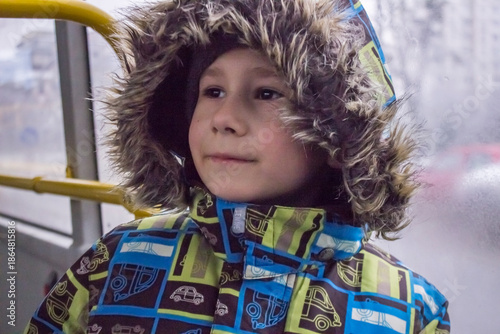 Little child looking through the window: a boy in a bright winter jacket with a faux fur hood and gloves. 