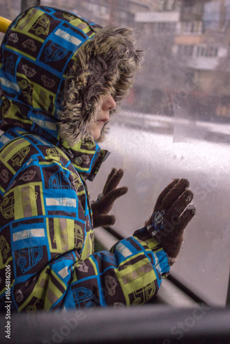 Little child looking through the window: a boy in a bright winter jacket with a faux fur hood and gloves. 