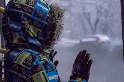Little child looking through the window: a boy in a bright winter jacket with a faux fur hood and gloves. 