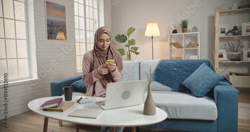 Muslim Woman in Hijab Using Smartphone While Working on Laptop at Home Modern Digital Lifestyle and Online Communication Concept