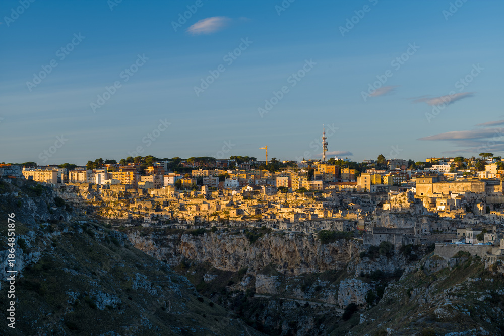 Fototapeta premium Soft golden light bathes the layered stone buildings of Matera, Italy, highlighting the dramatic cliffside setting beneath a clear blue sky with scattered clouds.