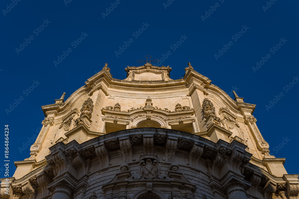Fototapeta premium Upward view of an ornate baroque church facade in Lecce, Italy, highlighted by warm sunlight against a deep blue sky. The intricate stonework and sculpted details showcase the architectural richness
