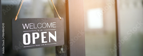 Black open sign with welcome text hanging on glass door of modern cafe, showing business opening hours and customer invitation. Vintage color tone style.