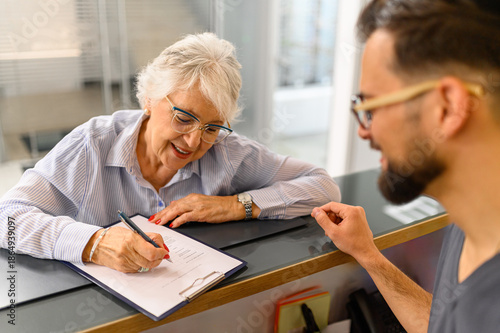 Photography Male receptionist looking at senior patient filling form of medical document at
