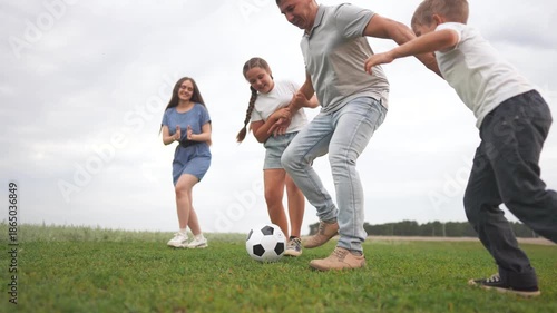 The girl, man, and boy play soccer on a grassy field while the girl runs towards the ball. The man kicks the soccer ball as the boy and girl interact nearby.