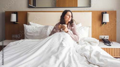 Calm woman sitting in a hotel bed with white bedding, holding a cup of coffee and looking away thoughtfully, cozy morning atmosphere, quiet moment of reflection, lifestyle and emotions concept