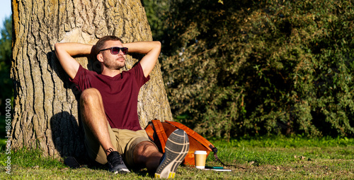 Urban man relaxing in city park, sitting near tree with hands behind head.