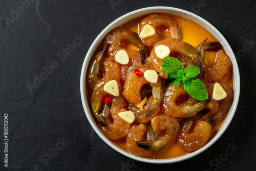Korean style soy marinated shrimp served with garlic slices, chili, and fresh herbs in a white bowl on dark background. Popular Korean seafood dish known as saeu jang, premium raw shrimp,