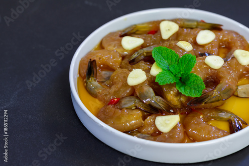 Korean style soy marinated shrimp served with garlic slices, chili, and fresh herbs in a white bowl on dark background. Popular Korean seafood dish known as saeu jang, premium raw shrimp,