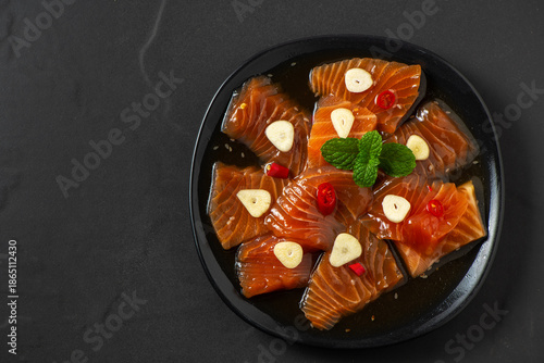 Top view Korean-style soy marinated salmon slices served with garlic, red chili, and herbs on a black plate. Popular Korean food known as salmon jang, premium seafood dish