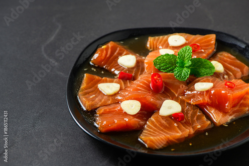 Korean-style soy marinated salmon slices served with garlic, red chili, and herbs on a black plate. Popular Korean food known as salmon jang, premium seafood dish