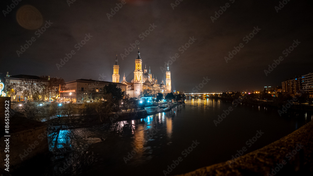Fototapeta premium Dynamic wide-angle night shot of the Basilica del Pilar in Zaragoza, with its illuminated towers and domes beautifully reflected in the flowing waters of the Ebro River during the Christmas season.