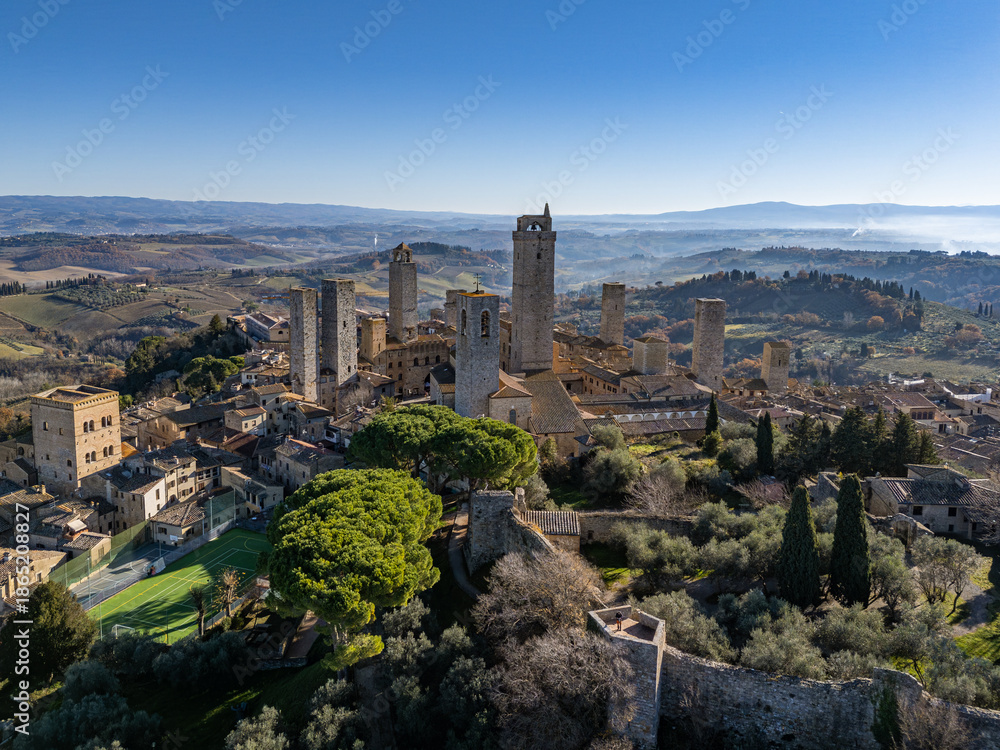 Fototapeta premium Aerial view of the medieval walled town of San Gimignano in Tuscany, featuring its iconic stone towers, ancient fortifications, and rolling hill landscape