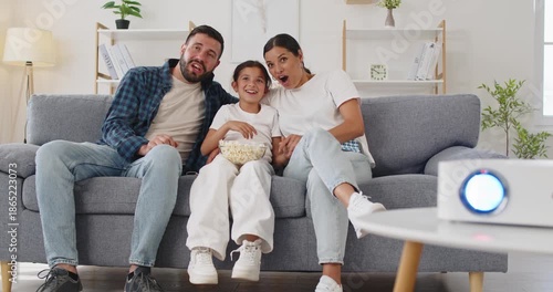 Family watching movie at home with projector. Parents and child sit on a cozy sofa couch in the living room, reacting with surprise to the big TV screen entertainment. Shared joy in home cinema.