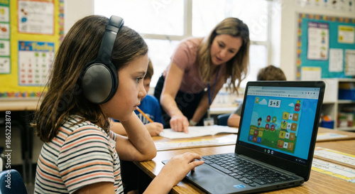 Girl sits at desk using laptop for math class while teacher helps in the background.