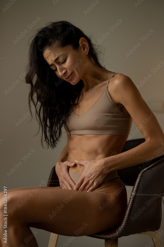 Naklejka premium Adult woman experiencing menstrual pain while sitting indoors, holding lower abdomen during period cramps, concept of women’s health, menstruation discomfort, hormonal cycle and female wellbeing.