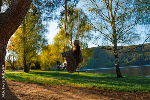 Woman in casual clothes and cap enjoying a wooden swing hung from a tree in a spring park by a calm lake, surrounded by lush green grass and trees with fresh foliage under warm evening light in Czech