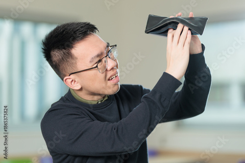 A disappointed Chinese student wearing glasses holds his empty wallet upside down showing no money inside, ideal for broke student, financial struggle, or budget crisis concepts.