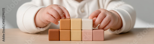 Baby hand stacking wooden block toy in warm neutral colors on wooden table, close up of child playing with educational cube, soft natural light creating calm mood
