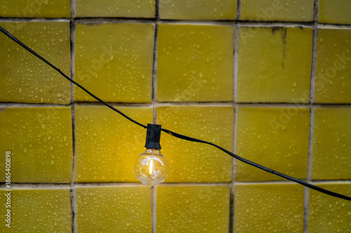 Close-up of a decorative outdoor string light illuminated against wet yellow ceramic tiles. Minimalist concept for energy, ideas, or cozy atmosphere