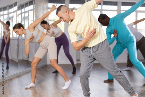 Positive expressive young man in casual sportswear learning modern street dancing during group class in choreographic studio