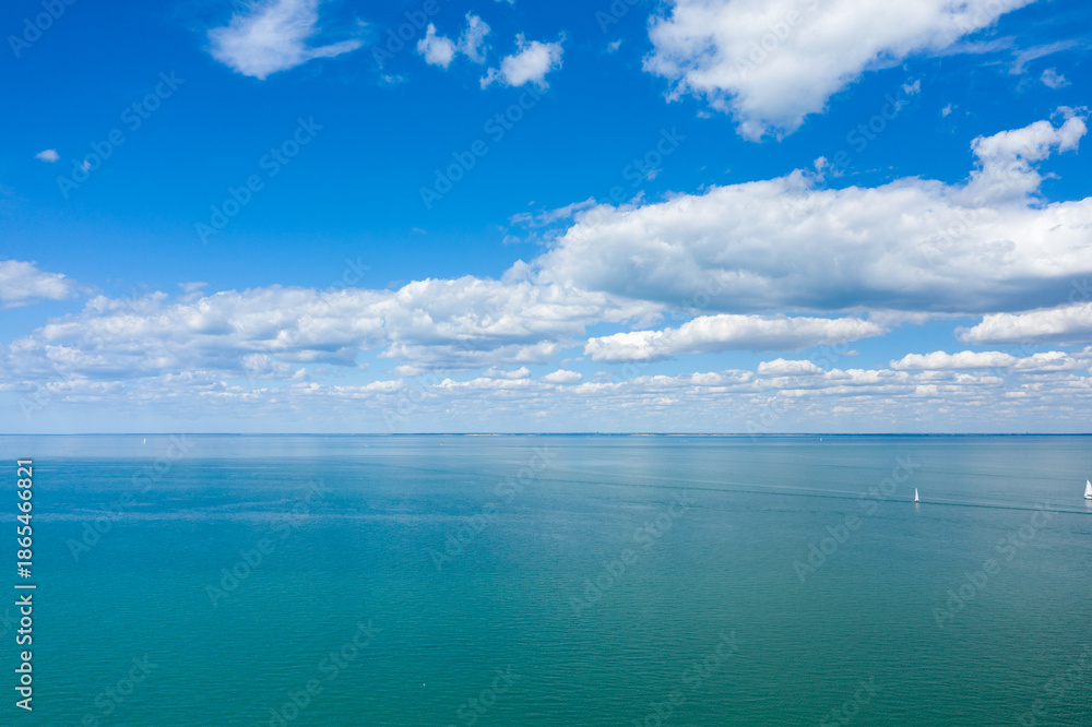Obraz premium Vast turquoise waters of the Atlantic Ocean stretch to the horizon beneath a sky filled with puffy white clouds. A few small sailboats dot the tranquil scene near Ile de Re, evoking a sense of space