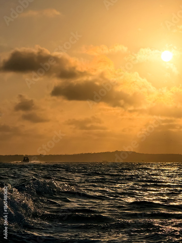 Photos Golden-hour sunset over the Taperoá Channel in Bahia, Brazil, with a motorboat wake in the foreground, choppy water, and distant boats on the horizon