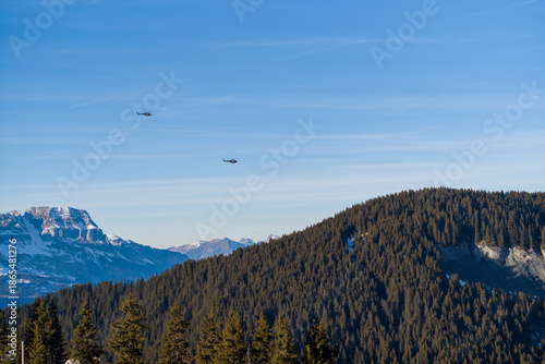 Valokuva Two helicopters soar over a densely wooded mountain ridge with snowy peaks in the distance, under a clear blue sky in the French Alps
