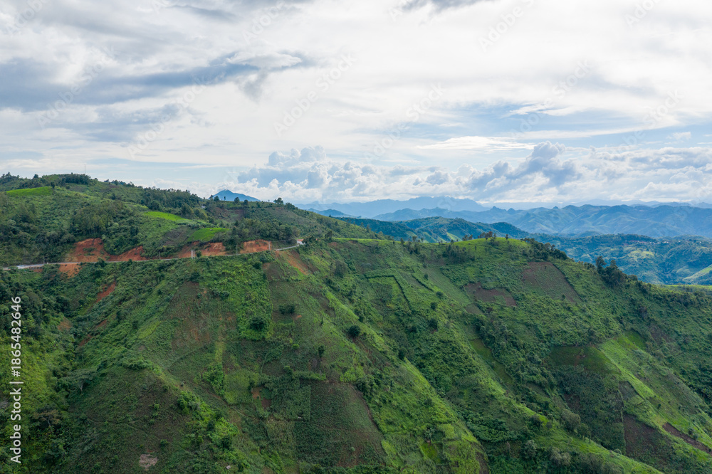 Naklejka premium A narrow mountain road curves along steep, lush green hillsides with distant blue ridges under a partly cloudy sky in the rural region between Phonsavan and Phou Khoun, Laos. The landscape features