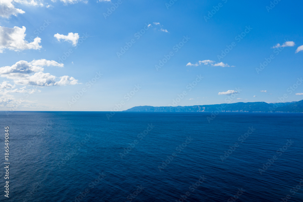 Obraz premium Vast expanse of deep blue Mediterranean water stretching toward a hazy mountainous coastline under a bright sky with scattered clouds near Orosei, Sardinia.
